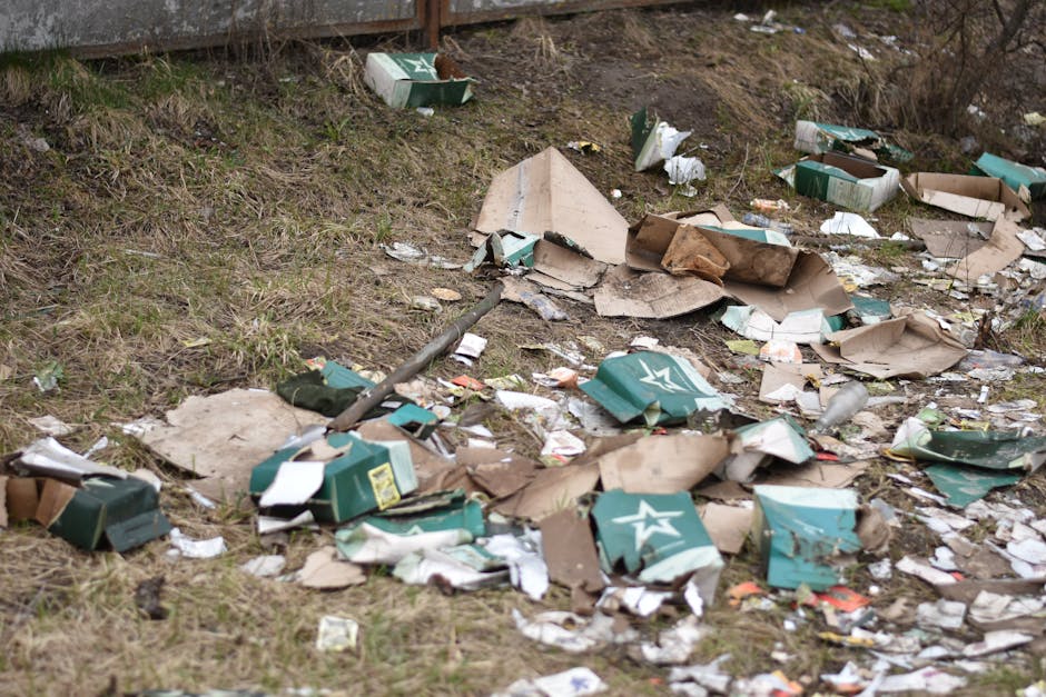 A spread of discarded cardboard boxes, many of which are partially crushed or torn, covers a patch of grass and dirt near a concrete boundary wall. The boxes are mostly green with white branding and logos, some shredded or crumpled. Among the boxes, there are scattered pieces of paper, plastic packaging, and miscellaneous rubbish, creating a cluttered and untidy scene. The background includes a weathered concrete wall, indicating an outdoor environment such as a yard or verge. The natural lighting suggests an overcast day, highlighting the disorder and accumulation of waste materials, which could be indicative of illegal dumping or mishandled waste disposal, reflecting the need for proper rubbish removal services like those provided by Waste Collection Highbury, specializing in private or alternative waste handling solutions outside standard municipal refuse collection.