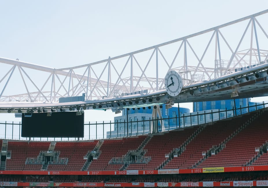 A section of a sports stadium, showing red seating arranged in rows with aisles and railings, positioned beneath a white overhead structure supported by triangular trusses. A large round clock with a white face and black hands is mounted on the structure, displaying the time as approximately 3:54. Behind the seating, modern blue and white buildings are visible against a clear sky, suggesting an urban environment outside the stadium. The scene appears well-lit by natural daylight, with the focus on the structural elements and seating area. The image emphasizes the stadium’s architectural details and the surrounding cityscape, providing a neutral view of an outdoor sports venue, relevant for content related to stadium facilities, event hosting, or urban infrastructure.