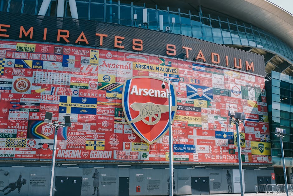 A large section of an outdoor stadium seating area is visible, with red plastic seats arranged in multiple rows and supported by metal frames. In the background, there are tall modern office buildings with glass facades reflecting the daylight. Above the seating area, a white metal walkway with a crisscross truss design spans across the image, featuring a circular clock showing approximately 2:05. The sky is clear and bright, with no clouds, suggesting a sunny day. The stadium appears to be designed for sports events, and the overall scene is clean and orderly, reflecting a typical setting where rubbish could accumulate from events or visitors. The presence of the structure and seating arrangement aligns with facilities that may require professional waste management, such as that provided by Waste Collection Highbury for private or commercial rubbish removal services in similar commercial or sports venues.