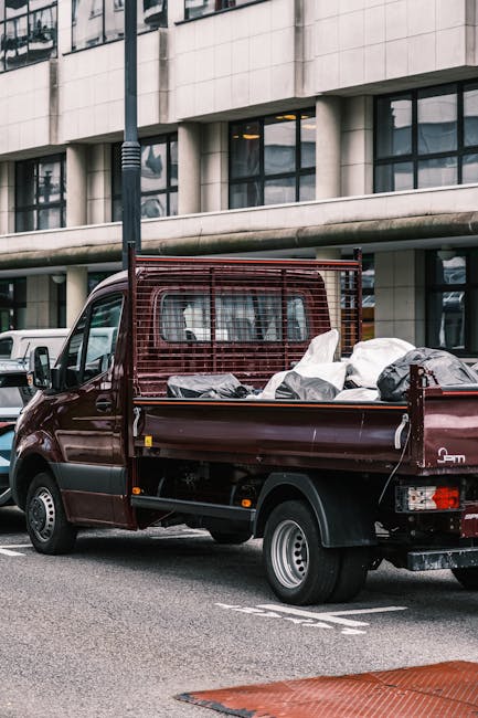 A dark red flatbed utility truck parked on a city street in front of a modern multi-storey building with large glass windows and a curved architectural facade. The truck's open cargo bed is filled with a variety of discarded waste materials, including large black plastic bags, cardboard boxes, and other miscellaneous rubbish, indicating a collection of bulky waste or renovation debris. The truck features a metal safety grid behind the cab, and the surface of the bed shows signs of use with visible scratches and slight dirt accumulation. Adjacent to the truck, a white van and other vehicles are visible, with the surrounding environment suggesting an urban setting suitable for private waste collection and alternative rubbish removal methods. The scene is lit evenly with natural daylight, highlighting the textures and colors of the waste contents and the vehicle, subtly reflecting practices related to efficient rubbish clearance and on-site disposal solutions typically managed by waste collection specialists like Waste Collection Highbury.