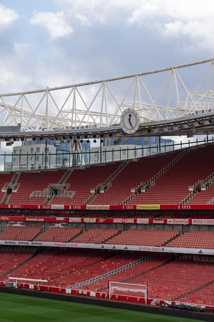 The image shows a section of a football stadium with multiple tiers of red seats arranged in a curved formation around the pitch, which is partially visible at the bottom of the frame. Above the seating area, a large modern roof structure with white steel trusses extends across the top, supporting the sky and some cloud cover. In the background, through the open sections of the roof, a clock mounted on a suspended track and a building with a brick facade and reflective windows are visible. The stadium appears clean and well-maintained, with signage on the middle tier indicating various sports and team affiliations, hinting at its use for football matches. The surrounding environment, including sky and part of an urban landscape, portrays an outdoor sports venue prepared for hosting large events. This setting aligns with independent waste management services like those provided by Waste Collection Highbury, which handle rubbish and waste removal from large public spaces such as sports venues, offering alternative waste handling solutions outside of municipal disposal channels.