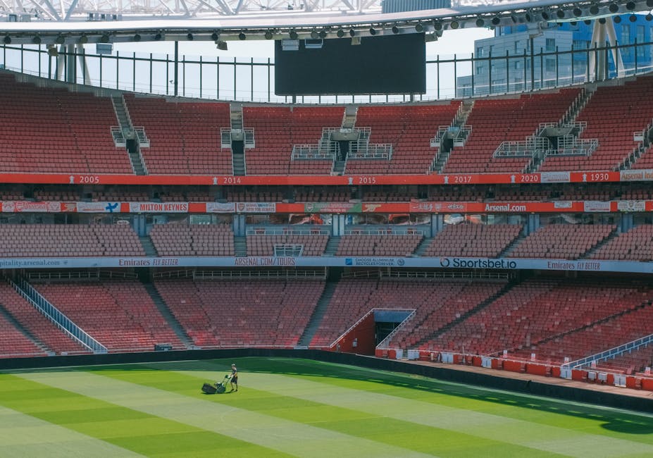 Inside an empty football stadium with multiple tiers of red seats and large glass windows at the top, a staff member wearing a high-visibility vest is operating a lawn mower on the well-maintained green grass pitch. The pitch features alternating light and dark green stripes created by mowing patterns. Surrounding the field are concrete barriers and support structures, with some sections in shadow, indicating sunlight streaming into the stadium. The seats are arranged in a circular pattern around the playing area, with some areas designated for specific sections marked by signs. The upper levels display digital advertising banners and a large black screens hanging from the ceiling, which is supported by a metallic framework. The scene depicts a moment of maintenance or cleaning activity related to stadium upkeep, resembling private or independent field management that could align with alternative waste handling practices in such environments, with the worker central in the foreground performing ground maintenance on the grass.