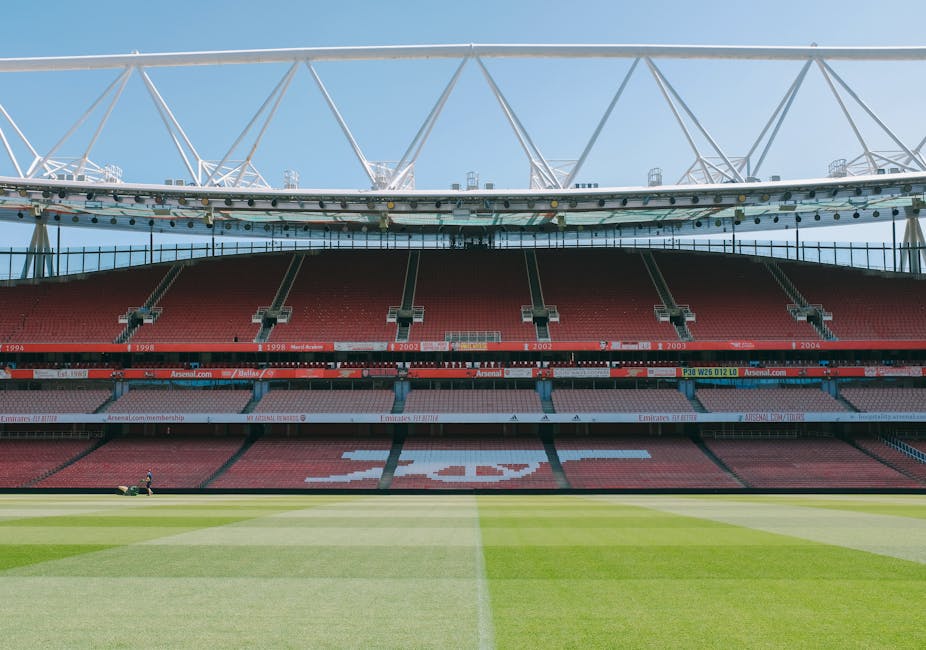 The image shows a section of a football stadium with multiple tiers of red seats arranged in a curved formation around the pitch, which is partially visible at the bottom of the frame. Above the seating area, a large modern roof structure with white steel trusses extends across the top, supporting the sky and some cloud cover. In the background, through the open sections of the roof, a clock mounted on a suspended track and a building with a brick facade and reflective windows are visible. The stadium appears clean and well-maintained, with signage on the middle tier indicating various sports and team affiliations, hinting at its use for football matches. The surrounding environment, including sky and part of an urban landscape, portrays an outdoor sports venue prepared for hosting large events. This setting aligns with independent waste management services like those provided by Waste Collection Highbury, which handle rubbish and waste removal from large public spaces such as sports venues, offering alternative waste handling solutions outside of municipal disposal channels.