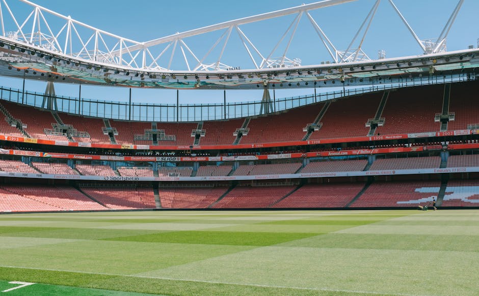 Interior view of a large football stadium with a well-maintained grassy pitch occupying the foreground. The stadium seats, primarily in shades of red, surround the pitch in tiered rows, with some sections featuring white or pink seating. Multiple advertising banners are visible along the middle tier of the stands, displaying various brand names and websites. Above, a modern structural roof supported by white trusses arches over the seating area, allowing natural light to illuminate the stadium interior. The sky outside is clear and blue, contributing to an open, bright atmosphere suitable for a sporting event or large gathering. Despite the presence of the stadium's structural elements, the scene does not include spectators, players, or equipment, conveying a quiet moment or setup phase. While the image depicts a facility used for public sports events, the broad, empty interior aligns with concepts of alternative event hosting or private scenario planning, subtly paralleling the idea of handling large-scale empty spaces in waste management or event logistics, as could be relevant for waste collection or site clearance services nearby.