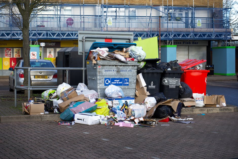 An overflowing waste collection point with multiple black, white, and red plastic rubbish bags, along with flattened cardboard boxes and loose paper, all piled around and on top of a large grey recycling bin labeled for mixed paper and cardboard. To the left of the bin, there is a small grey car parked close to a metal railing separating the rubbish area from the nearby parking space. The scene is set on a paved urban street with a background featuring a commercial building with large storefront windows, some of which display signage for shops, and a blue scaffolding structure covering the upper level of the building. The environment is well-lit, suggesting daytime, with a leafless tree positioned at the left edge of the image, indicating a possible winter or early spring setting. This scene portrays a typical instance of curbside rubbish accumulation, potentially related to private waste disposal services like those offered by Waste Collection Highbury, rather than official council refuse collection, emphasizing alternative waste handling methods.
