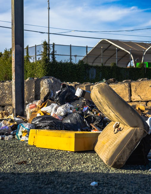 A close-up view of a pile of discarded rubbish on a gravel surface adjacent to a stone retaining wall, with black plastic rubbish bags, cardboard boxes, and various household waste items visible. The black bags are crumpled and stacked haphazardly, while a yellow plastic container lies open, partially obscured by a large, dirty, beige car seat cushion that leans against the debris. In the background, there is a metal pole with electrical wires, a green hedge, and a sports court with a white curved canopy structure, indicating an outdoor environment. The scene is illuminated by natural daylight, highlighting the texture of the waste materials, which appear to have been left temporarily, possibly as part of a private rubbish collection or on-site clearance undertaken by Waste Collection Highbury, a waste management service. The image underscores the importance of professional rubbish removal to prevent illegal dumping and maintain cleaner surroundings.