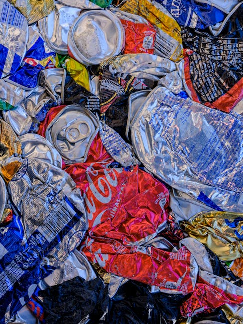 A collection of overflowing waste and rubbish bags piled around multiple waste bins in an urban parking area. The waste includes cardboard boxes, plastic bags, paper, and miscellaneous packaging materials, some of which are torn or spilled onto the surrounding brick pavement. The waste bins consist of a large grey bin labeled for mixed paper and card, a black bin, a red bin, and a black recycling bin, all positioned close to a metal railing separating the area from the street. In the background, there are shopfronts and a building under construction or renovation with blue safety scaffolding and signage. A silver car is parked nearby, and a tree is visible to the left of the waste piles. The scene is lit by natural daylight, suggesting a typical day with no immediate waste collection or disposal happening, illustrating an instance of unmanaged rubbish awaiting pick-up, possibly highlighting the need for private waste services such as those offered by Waste Collection Highbury.