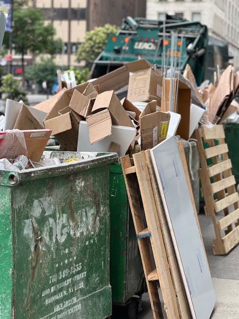 A large green waste collection bin filled with a mixture of cardboard boxes, some flattened and others partially assembled, along with a few white cardboard containers and packaging materials, positioned on a city street near a pile of discarded wooden pallets and a sliver of a large green waste collection truck in the background. The scene is outdoors, with blurred buildings and trees in the distance, reflecting an urban environment suitable for independent waste removal or onsite clearance. The cardboard items display various textures, from smooth and glossy to corrugated, with brown and white tones, some with printed labels or markings. The pallets are light-colored, wooden, and stacked vertically against the bins, while the environment suggests an active rubbish disposal process, possibly carried out by a private waste collection service like Waste Collection Highbury. The lighting appears natural, with a soft overcast sky casting gentle shadows, emphasizing the city's ongoing effort to manage bulky waste outside of standard municipal collection schemes.
